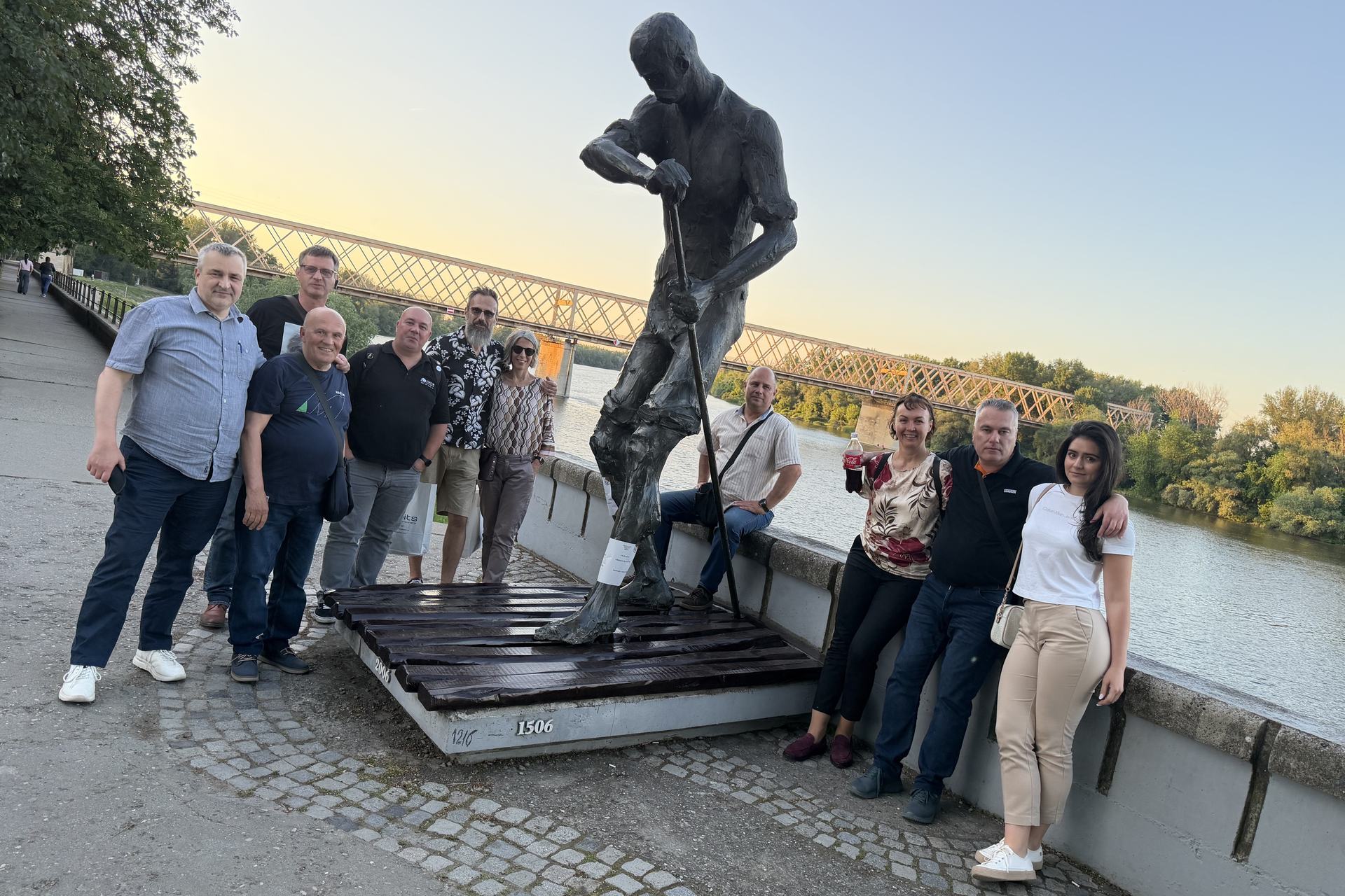 The eFTI Connect team and workshop guests beside the bronze raftsman on the Tisa embankment in Senta, with the old railway bridge behind them at sunset